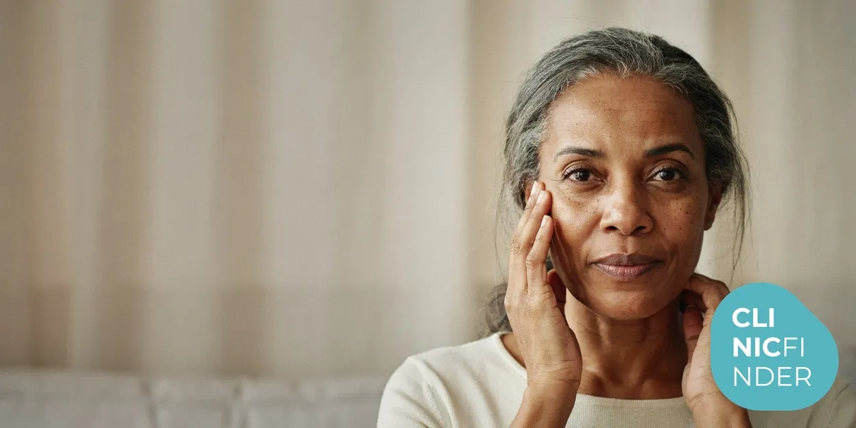 A Black woman in her fifties with radiant, healthy skin gently touching her cheek and jawline in soft natural daylight, reflecting the focus of non-surgical anti-ageing care