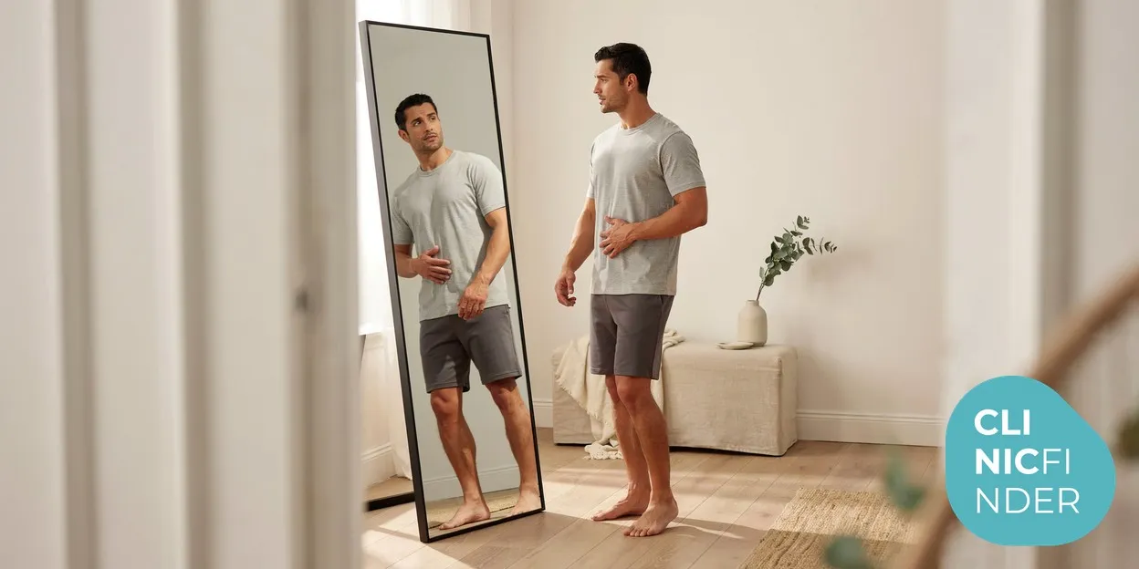 A man standing side-on in soft daylight, calmly assessing his profile and midsection in a full-length mirror while deciding between body-contouring options