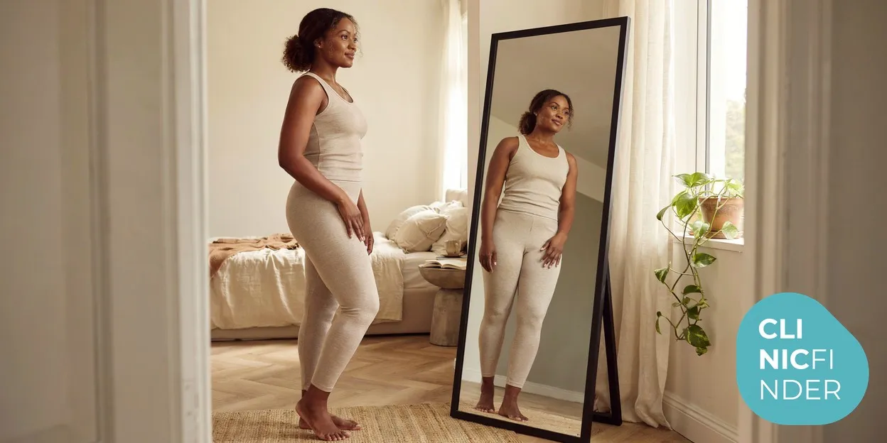 A woman standing side-on assessing her waist and flank silhouette in a full-length mirror in a bright room