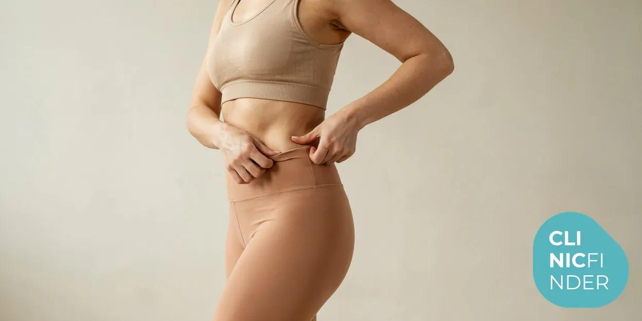 A woman in soft neutral activewear gently pinching a small pocket of fat at her waistline in warm natural daylight, illustrating stubborn pinchable fat