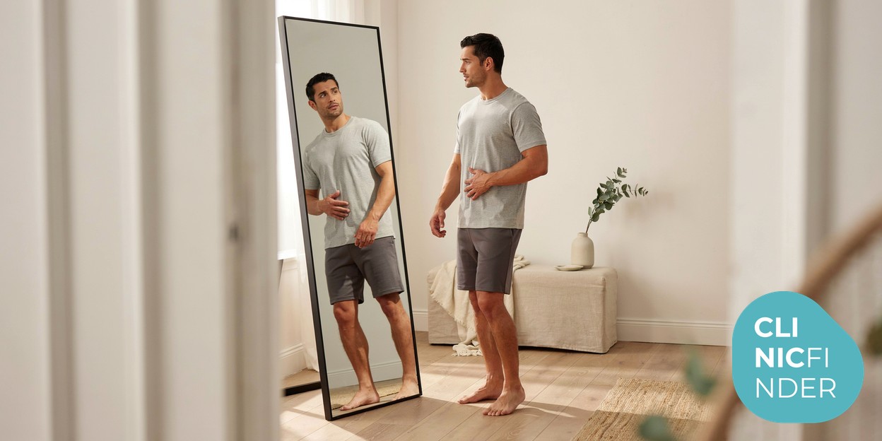 A man standing side-on in soft daylight, calmly assessing his profile and midsection in a full-length mirror while deciding between body-contouring options